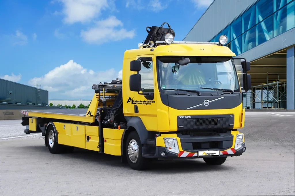 Żółta laweta Volvo FL z logo "LiderAssistance" zaparkowana przed nowoczesnym budynkiem. Pojazd jest wyposażony w reflektory dachowe i specjalistyczne narzędzia do transportu uszkodzonych pojazdów.